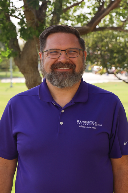 A white man wearing glasses and a purple polo smiles in front of a tree.