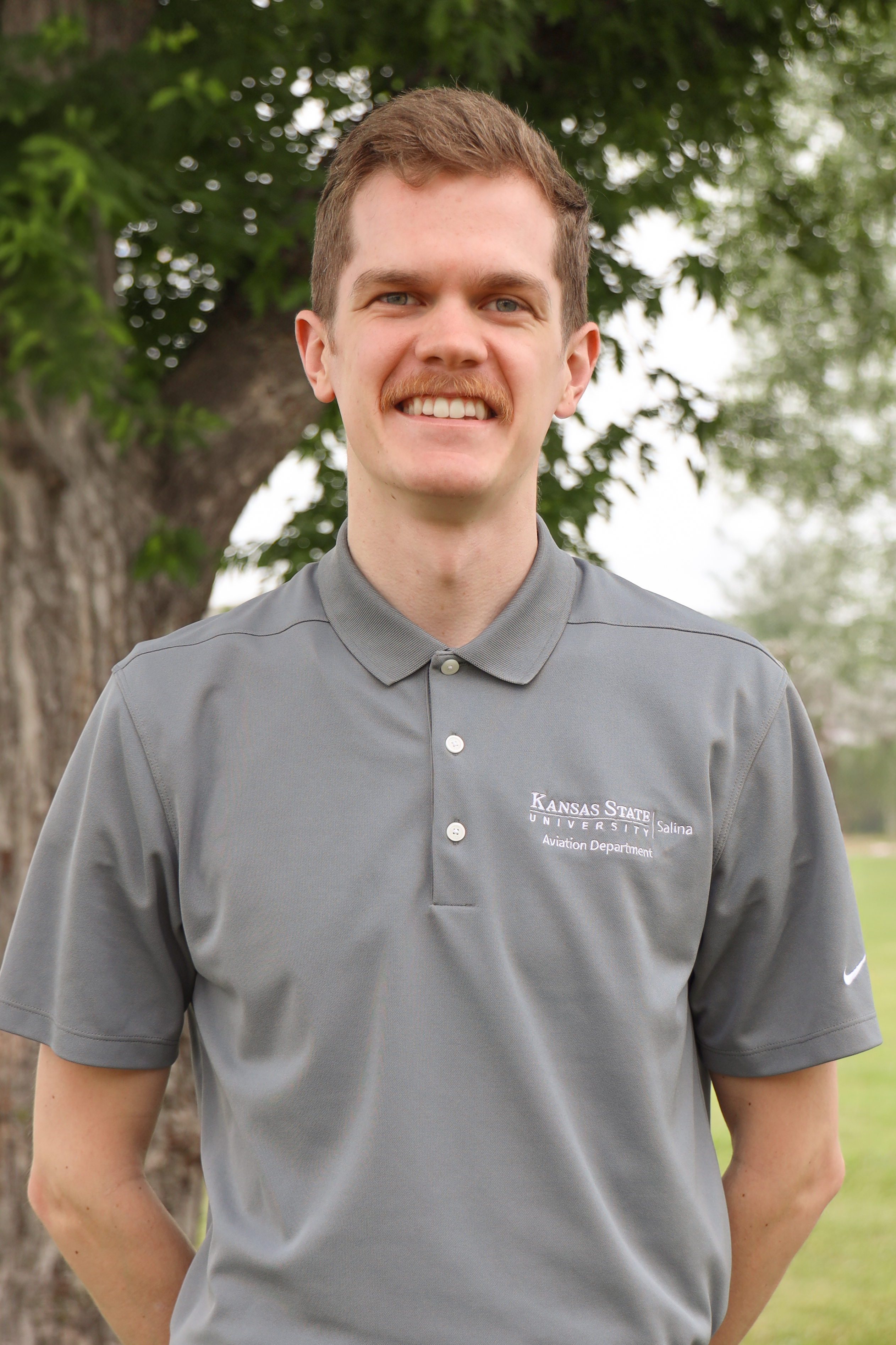 A white man with brown hair wearing a light gray polo smiles in front of a tree.