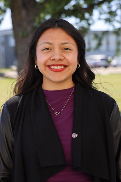 A woman with dark hair wearing a purple shirt and black cardigan smiles in front of a tree.