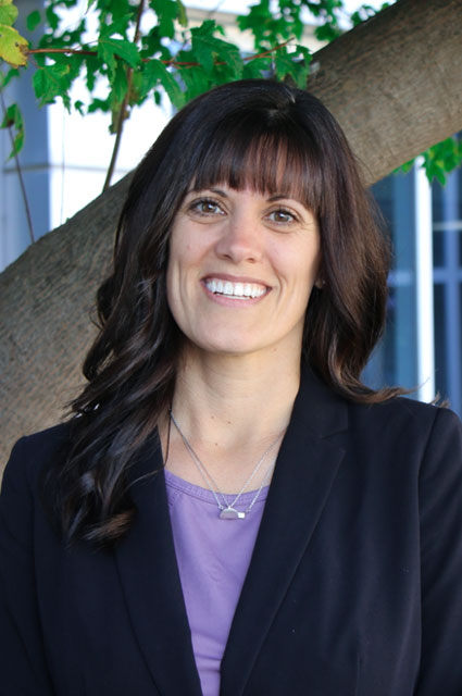 A woman with dark hair and bangs smiles in front of a tree.