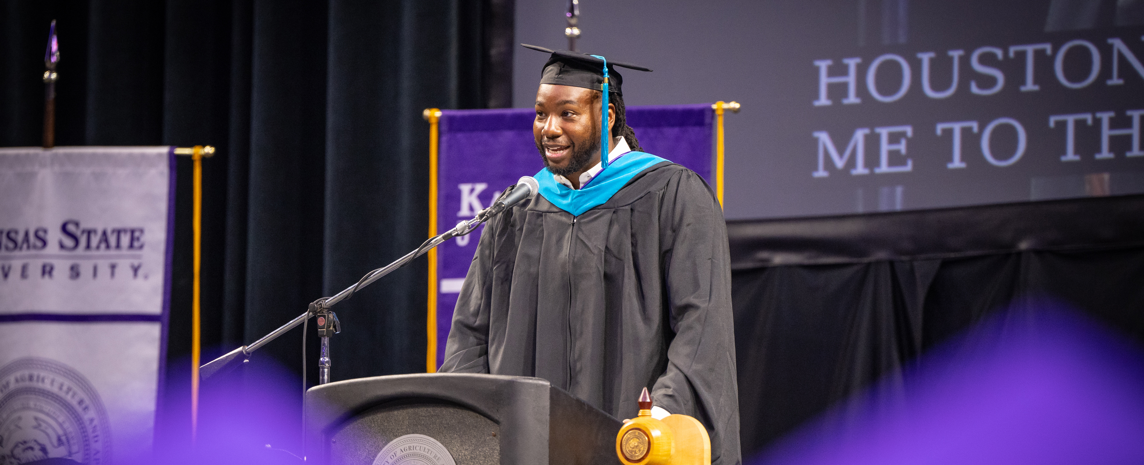 A Black man in graduation regalia speaks into a microphone in front of university flags.