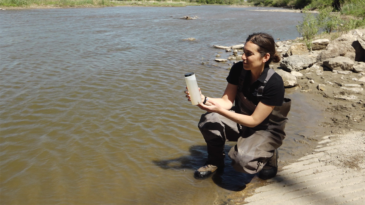 A woman in a black t-shirt and waders holds a container of water collected from the lake she's kneeling in.