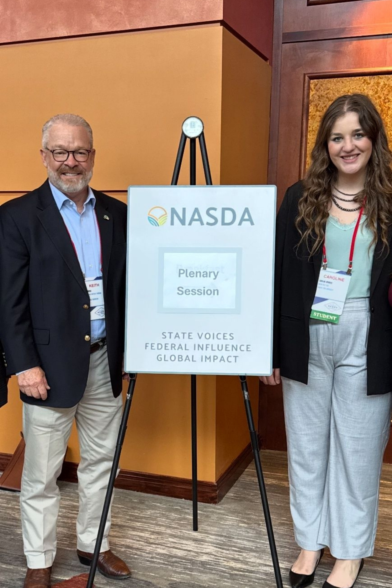 Wingert poses next to a conference signage poster during the NASDA annual meeting. The poster has the NASDA logo, and, below, "Plenary Session."