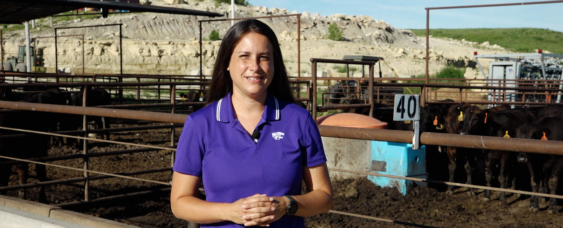 A woman with long brown hair wearing a purple polo stands in front of a cattle pen.