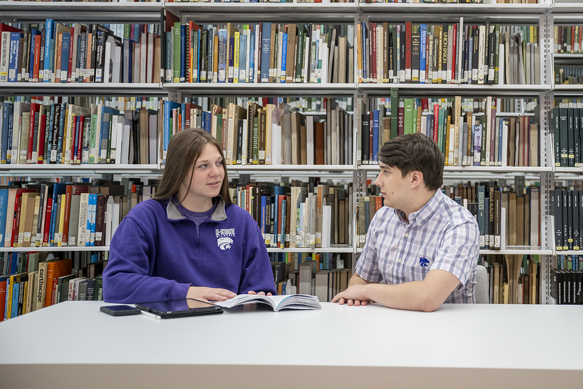 Two college architecture students sit at a table in a library and hold a discussion.