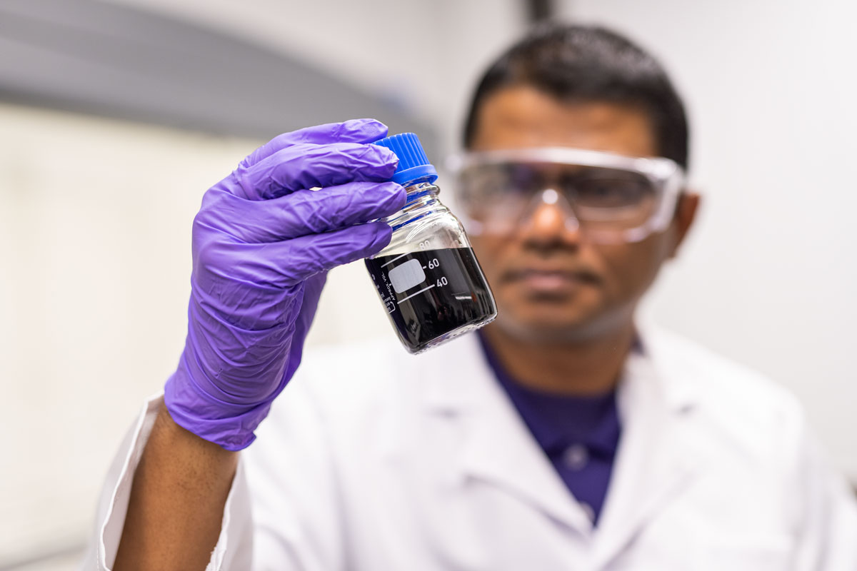 A person with a white lab coat, protective eyewear and purple gloves examines a glass jar of dark liquid.