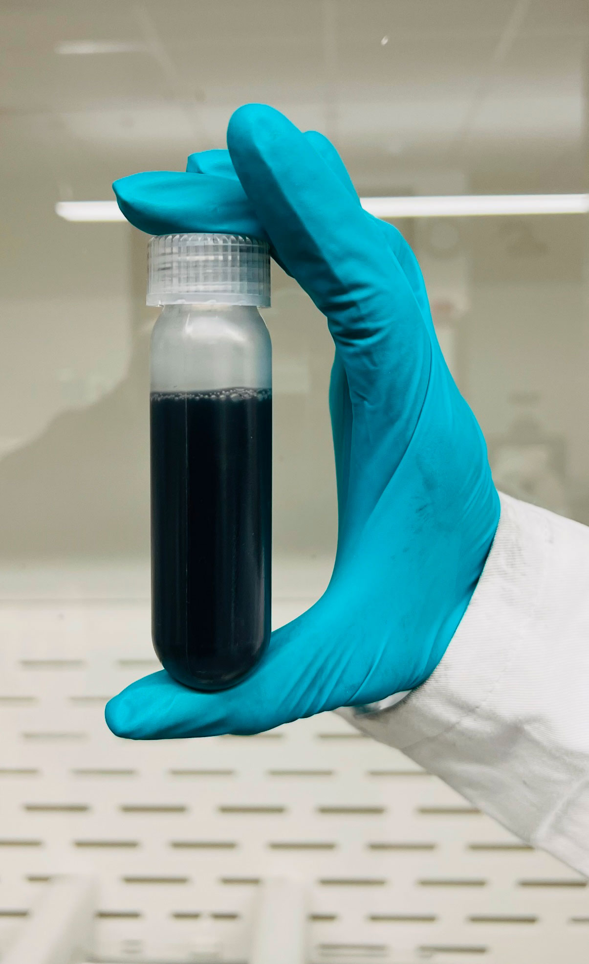 A hand with blue gloves holds a container of a dark liquid material.