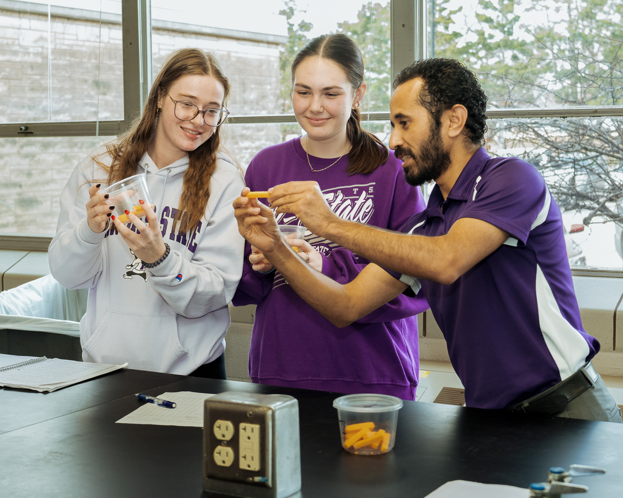 Three people, two female students and a male professor, stand at a black counter with windows behind them examining cheese.