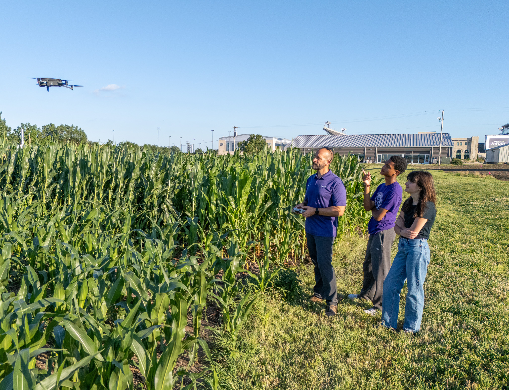 Three people stand outside in a green cornfield and operate a small drone that is hovering above the corn.