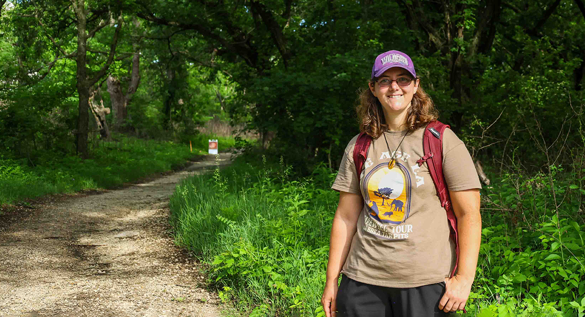 A doctoral student in a t-shirt, backpack and ballcap poses for a portrait on a wooded prairie trail.