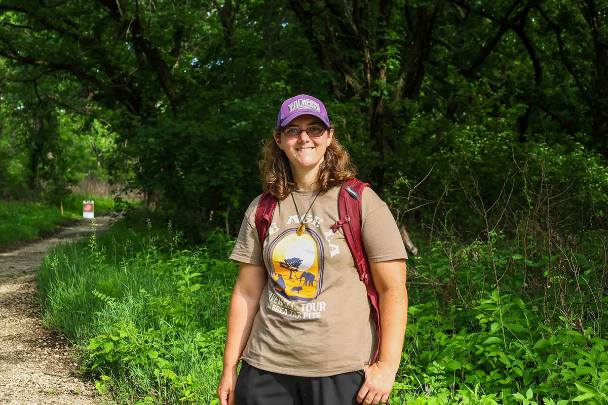 A doctoral student in a brown T-shirt, red backpack and purple hat poses for a portrait on a trail in a wooded prairie.
