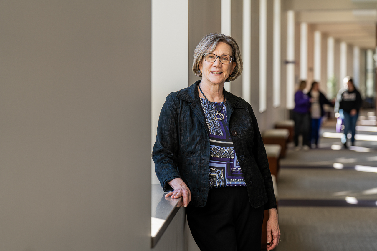Kerri Day Keller stands in business casual attire and poses for a professional portrait in a long hallway with narrow vertical windows.  