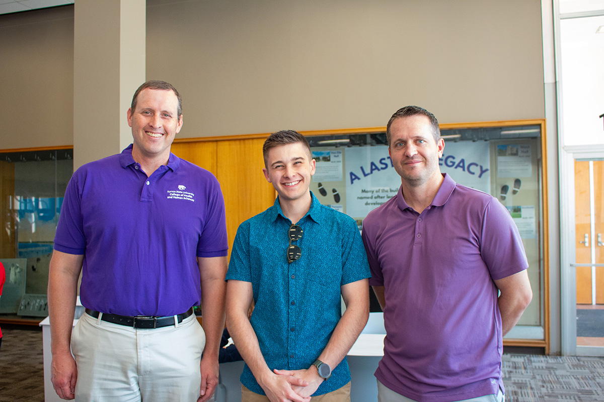 Three men pose in building lobby with man on left wearing purple polo shirt with logo and khaki pants, man in center wearing teal patterned shirt with sunglasses on collar, and man on right wearing light purple polo.