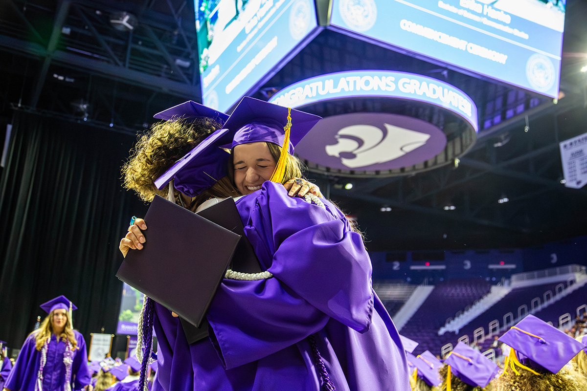 Three graduates in purple caps and gowns embrace on the arena floor with one holding diploma cover; gold tassels visible on mortarboards. Large video screen displaying "CONGRATULATIONS" and Powercat logo hangs above with arena seating and other purple-clad graduates visible in background.
