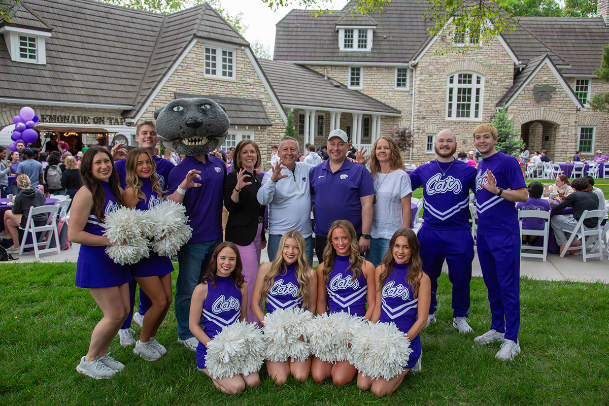 Willie the Wildcat mascot poses with a group of cheerleaders in purple uniforms holding silver pom-poms and staff members in purple and casual clothing on the lawn, with four cheerleaders kneeling in front row. Stone residential buildings with dormers, green trees, purple balloons, and white folding chairs visible in background under partly cloudy sky.
