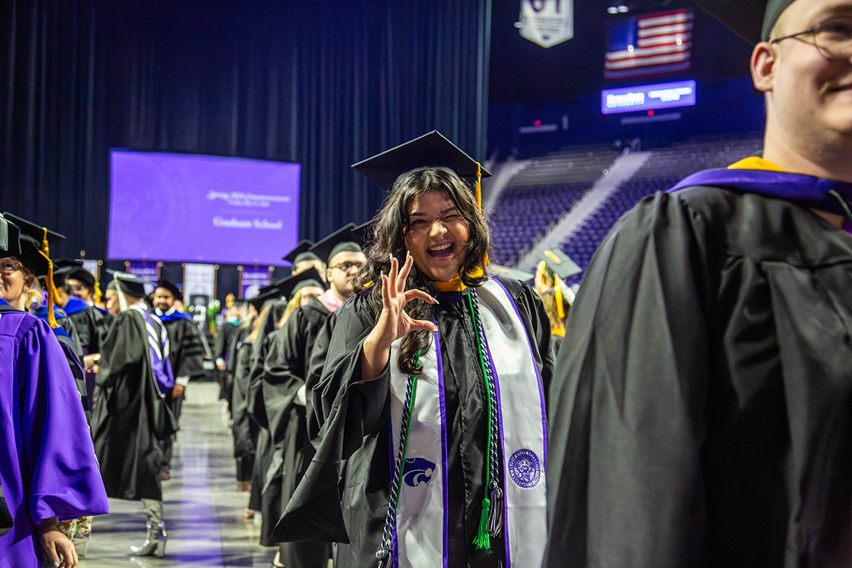 Graduate in black cap and gown throws up the Wildcat hand sign at the camera while walking across the arena floor wearing a white stole with green and purple cords and K-State medallion.