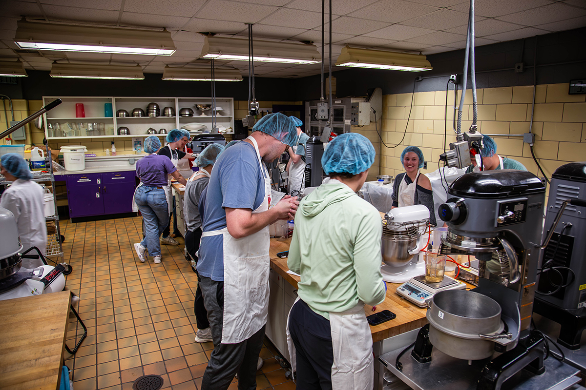 A group of college students wearing hairnets and aprons work in a college's baking laboratory.