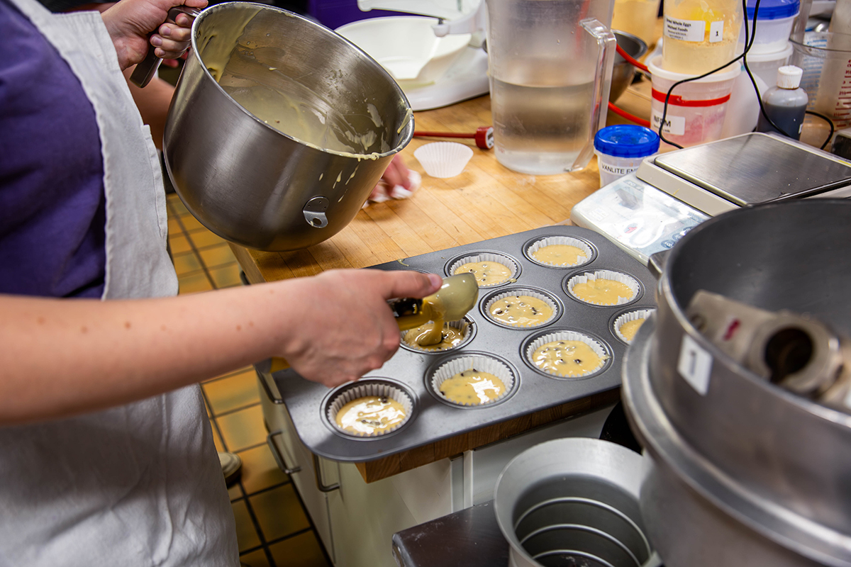 A student scoops globs of muffin dough into an aluminum muffin tin.