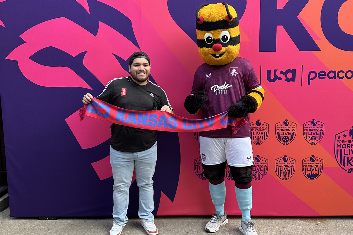 A man poses with a soccer mascot and both hold up a banner that says KANSAS CITY.