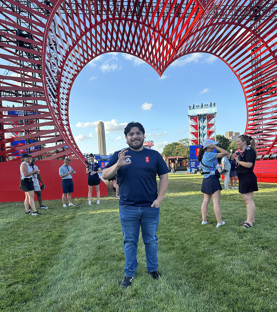 A man with dark hair and a beard stands in front of a large red heart gate structure at a fan festival for the World Cup.
