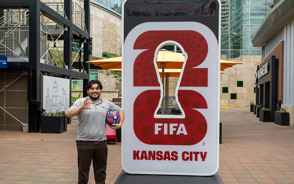 A man with dark hair and a beard holds a decorative soccer ball next to a FIFA Kansas City sign in an outdoor plaza.