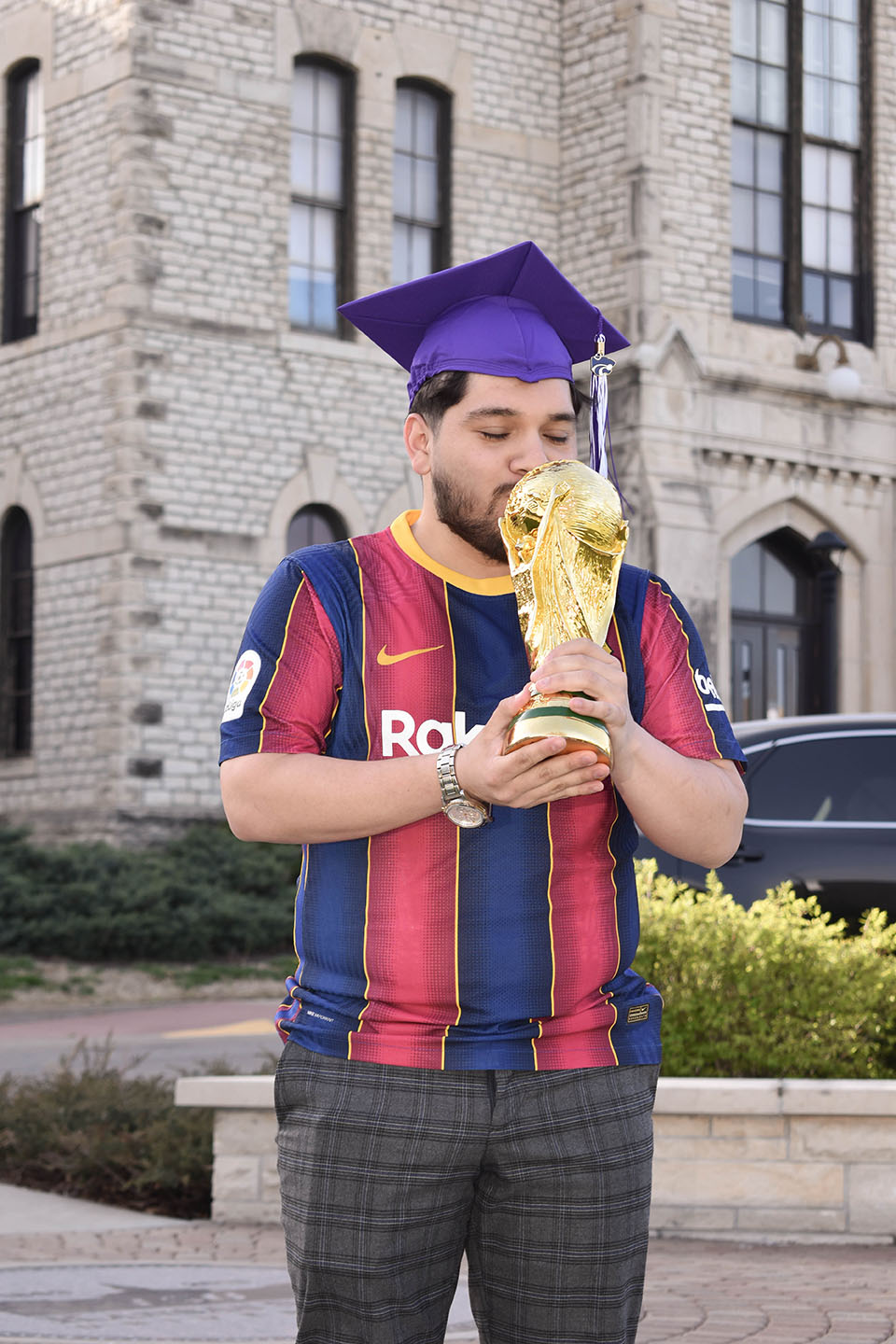 A man wearing a purple graduation cap wears a blue and red striped jersey and kisses a replica of the World Cup trophy.