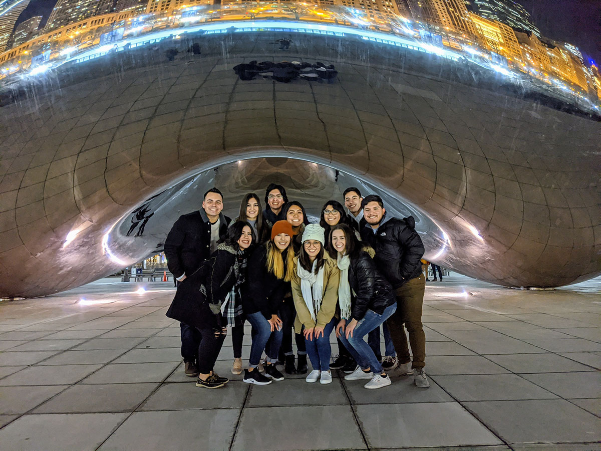 A group of ten students dressed in warm clothing stands below Cloudgate in Chicago.