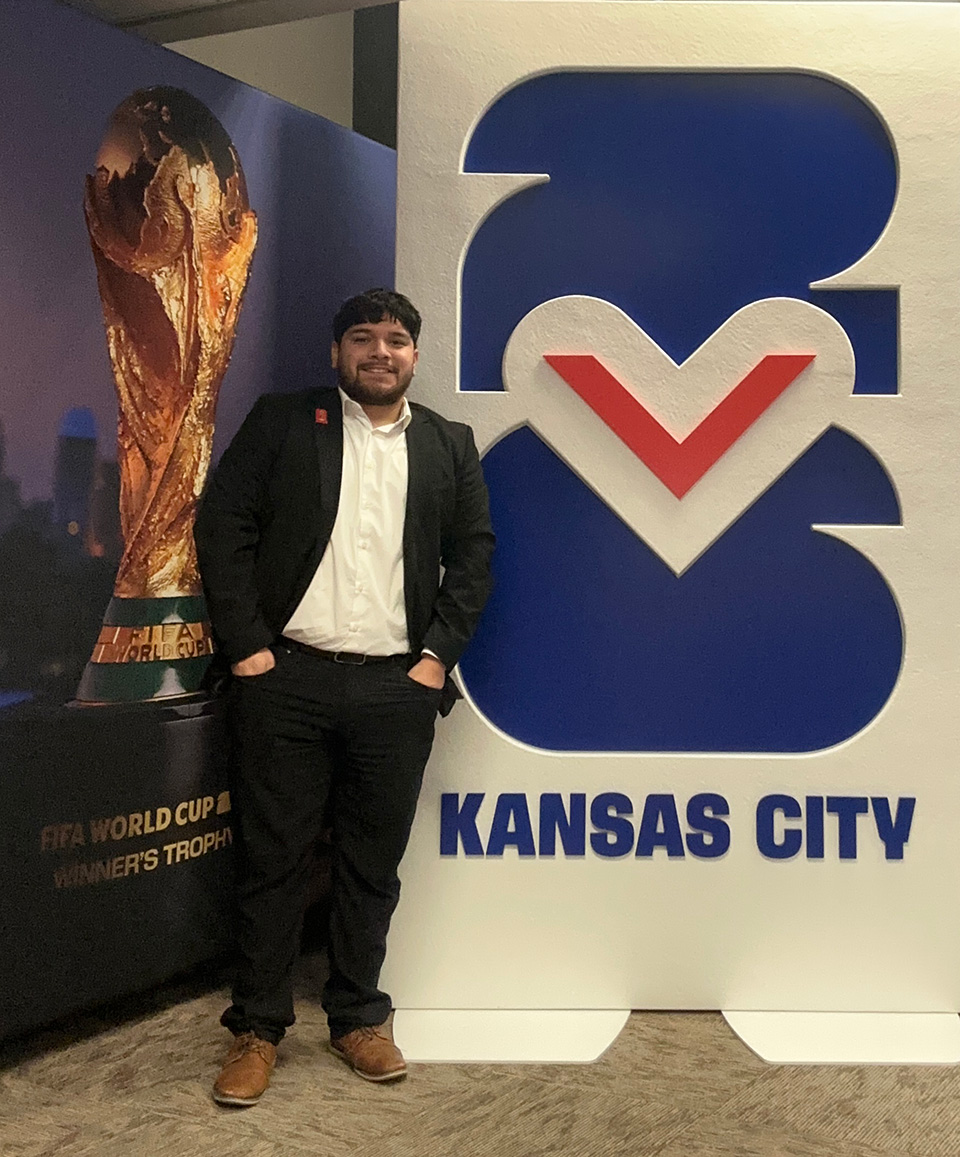 A man wearing a black suit jacket and pants stands between two signs for the FIFA World Cup in Kansas City.