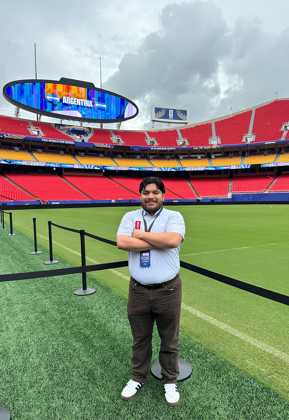 A man with dark hair and a short beard stands on a soccer field with red and yellow stadium seating in the background.