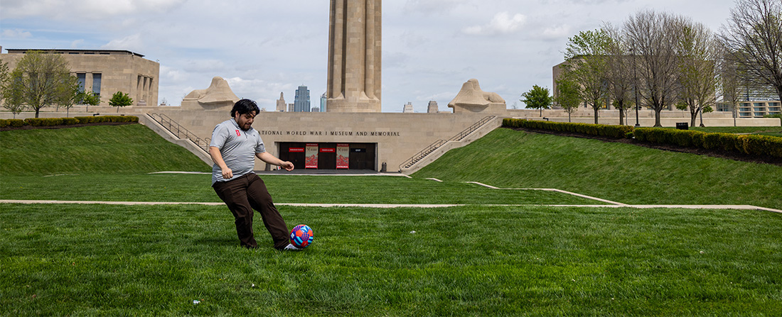 A man wearing dark pants and a gray polo kicks a colorful soccer ball on a field in front of a museum.