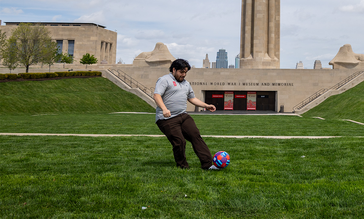 A man wearing dark pants and a gray polo kicks a colorful soccer ball on a field in front of a museum.