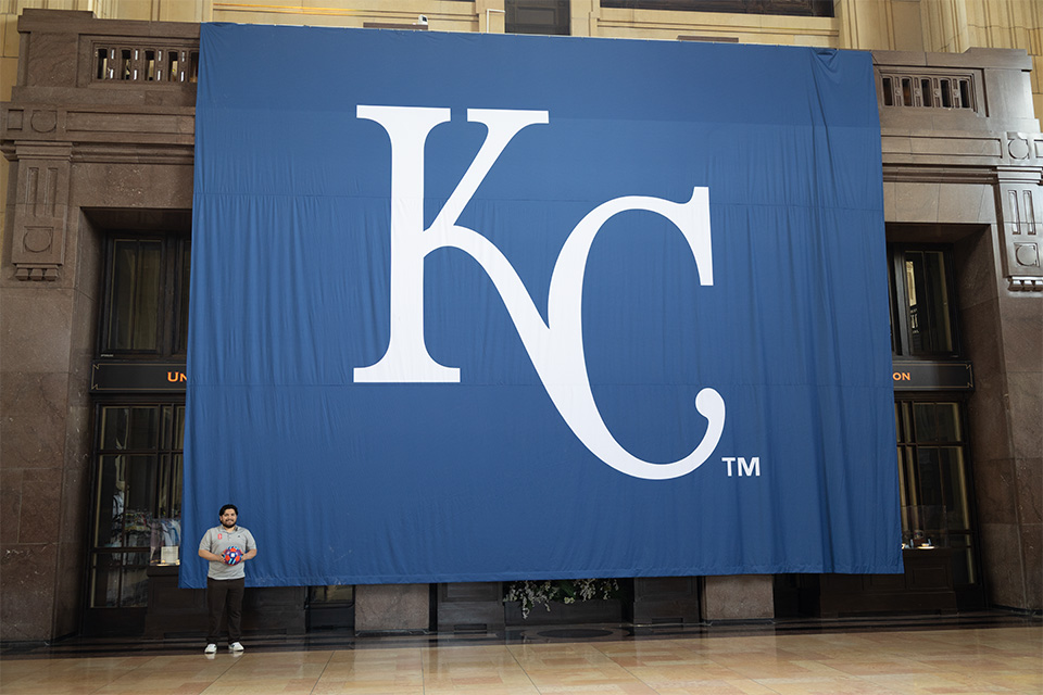 A man stands at the bottom left corner of a giant blue KC banner.