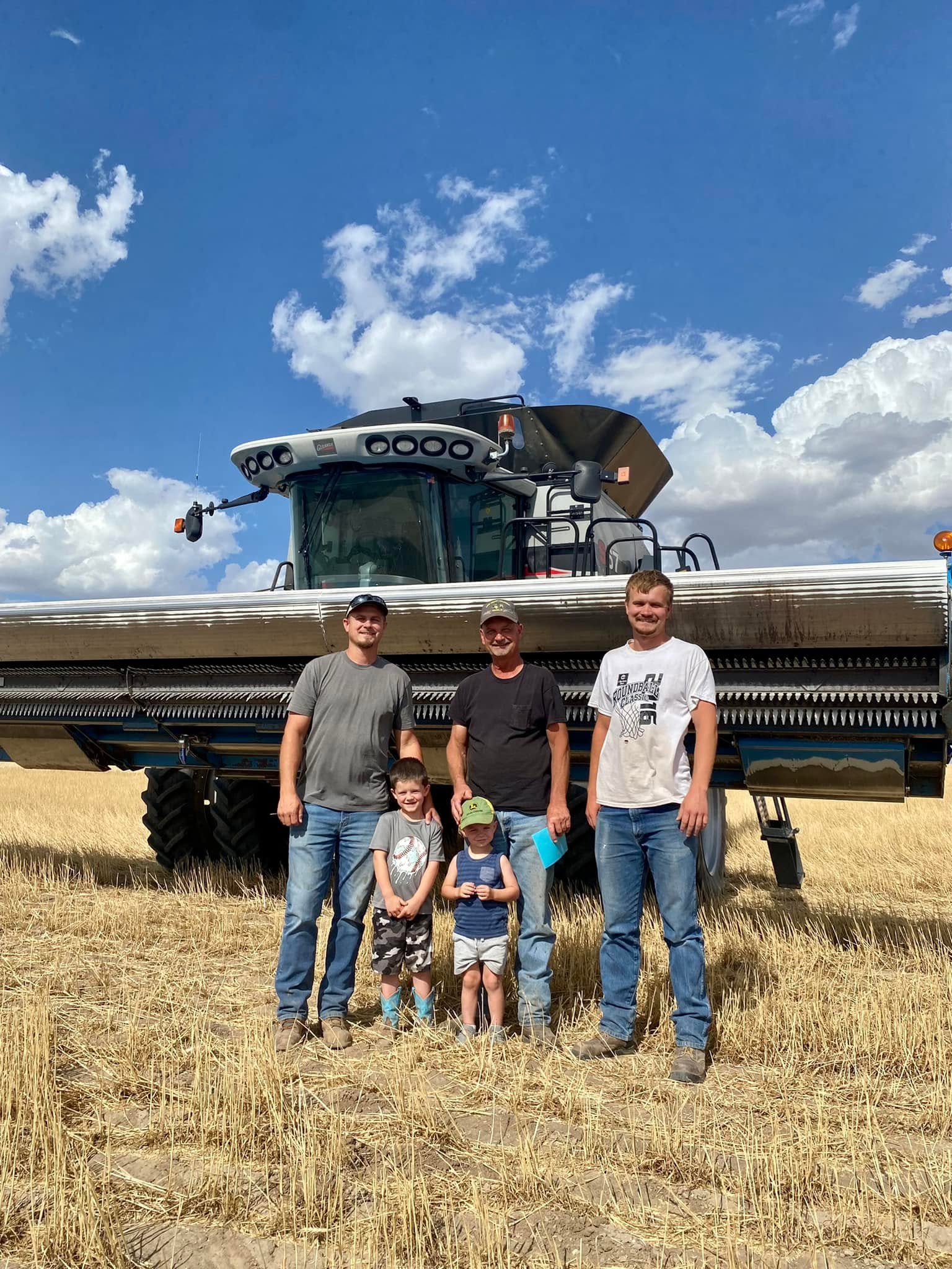 Three men and two kids stand in a field in front of a silver combine and a blue sky.