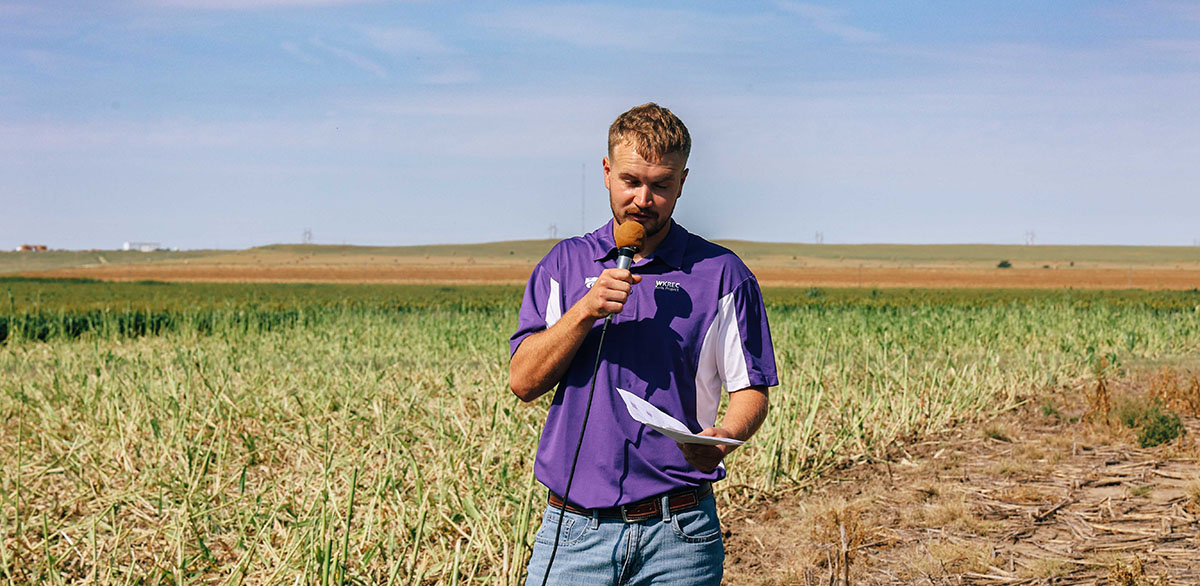 A man in a purple polo holds a microphone and reads from a paper in a field with blue sky behind him.