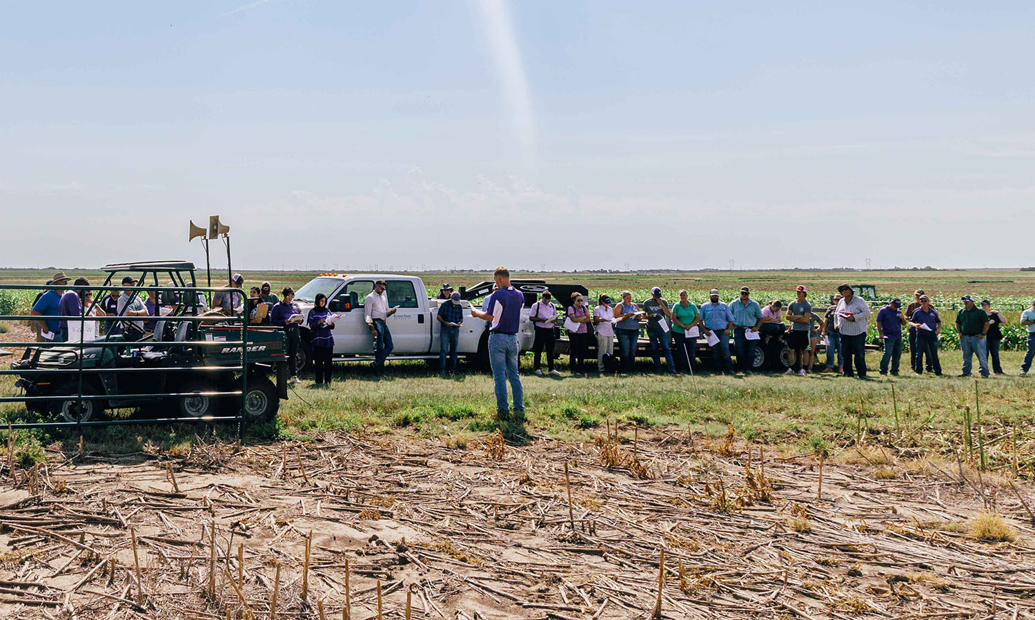 A man in a purple polo stands and presents to a large group of producers in a field