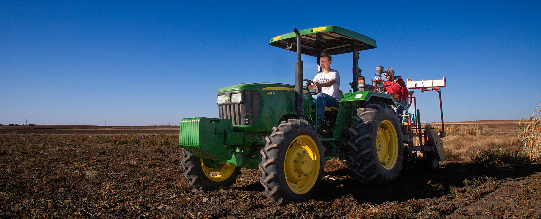 Two men ride a tractor in a brown field with a blue sky in the background.
