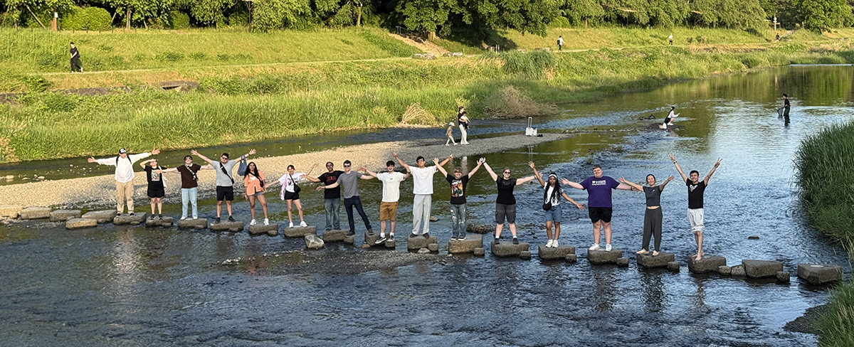 A group of people span a river on stepping stones. Most are raising their arms.