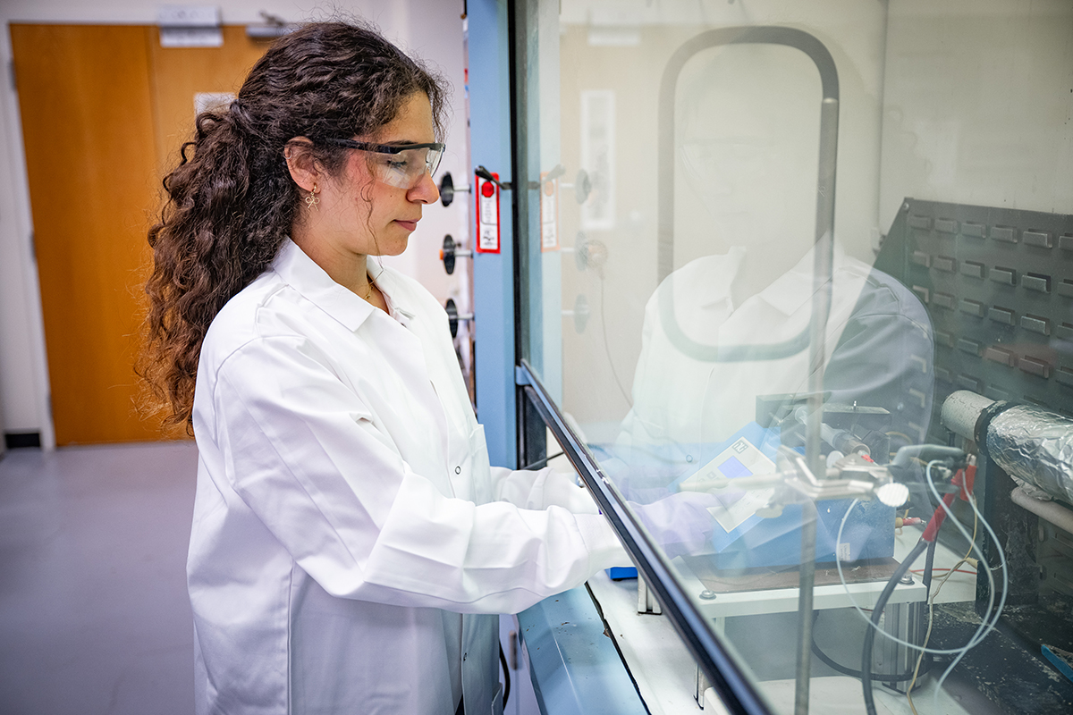 A female graduate student in a lab coat and protective glasses works with a piece of lab equipment under a hood.