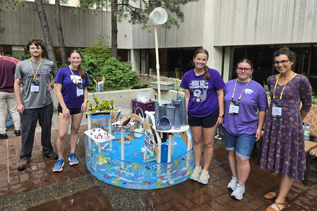 A group of college students in warm weather clothing and their advisor pose for a group photo next to a fountain they engineered in the middle of a plastic kiddie pool.