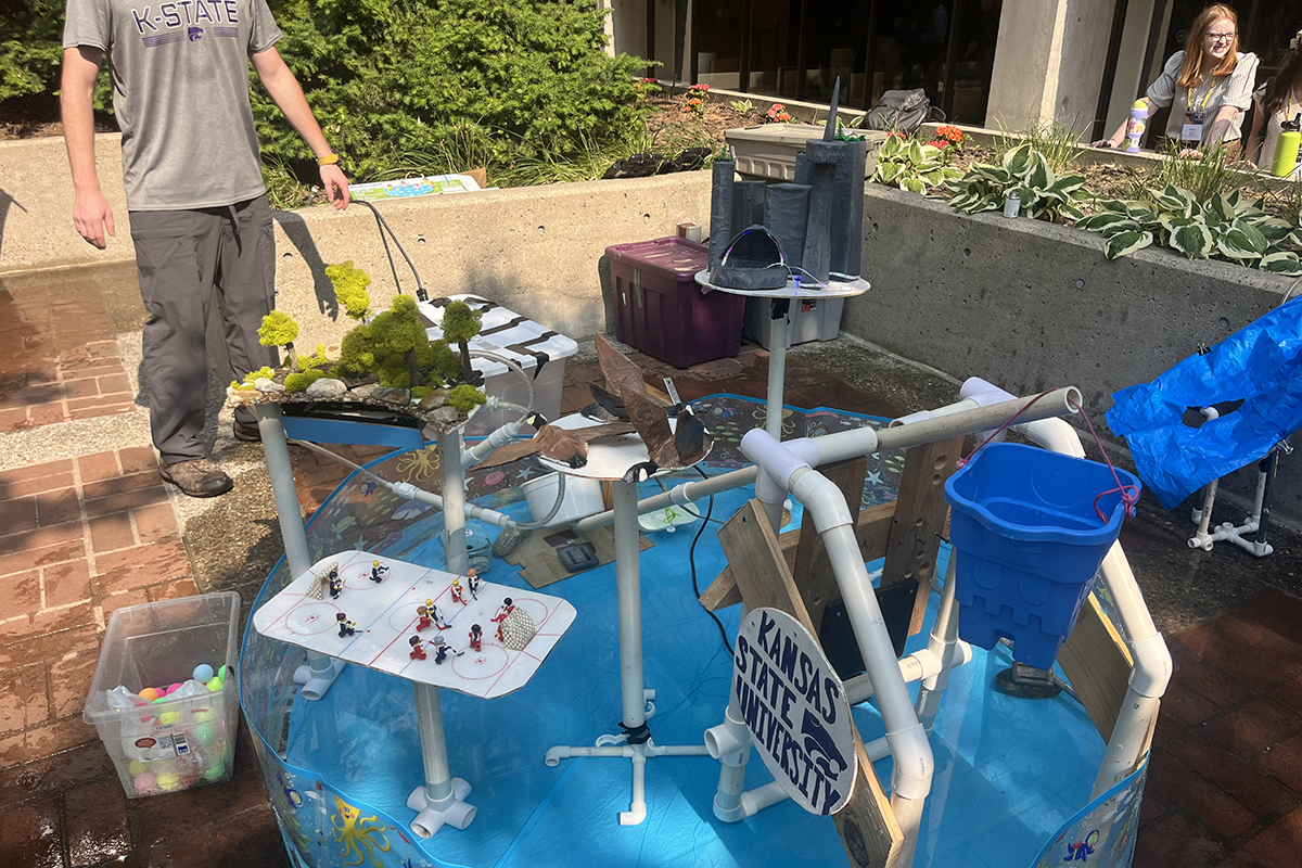 A contraption of PVC pipes, a miniature garden, a mini stone castle and other items stand on pipes in a plastic kiddie pool in an outdoor courtyard.