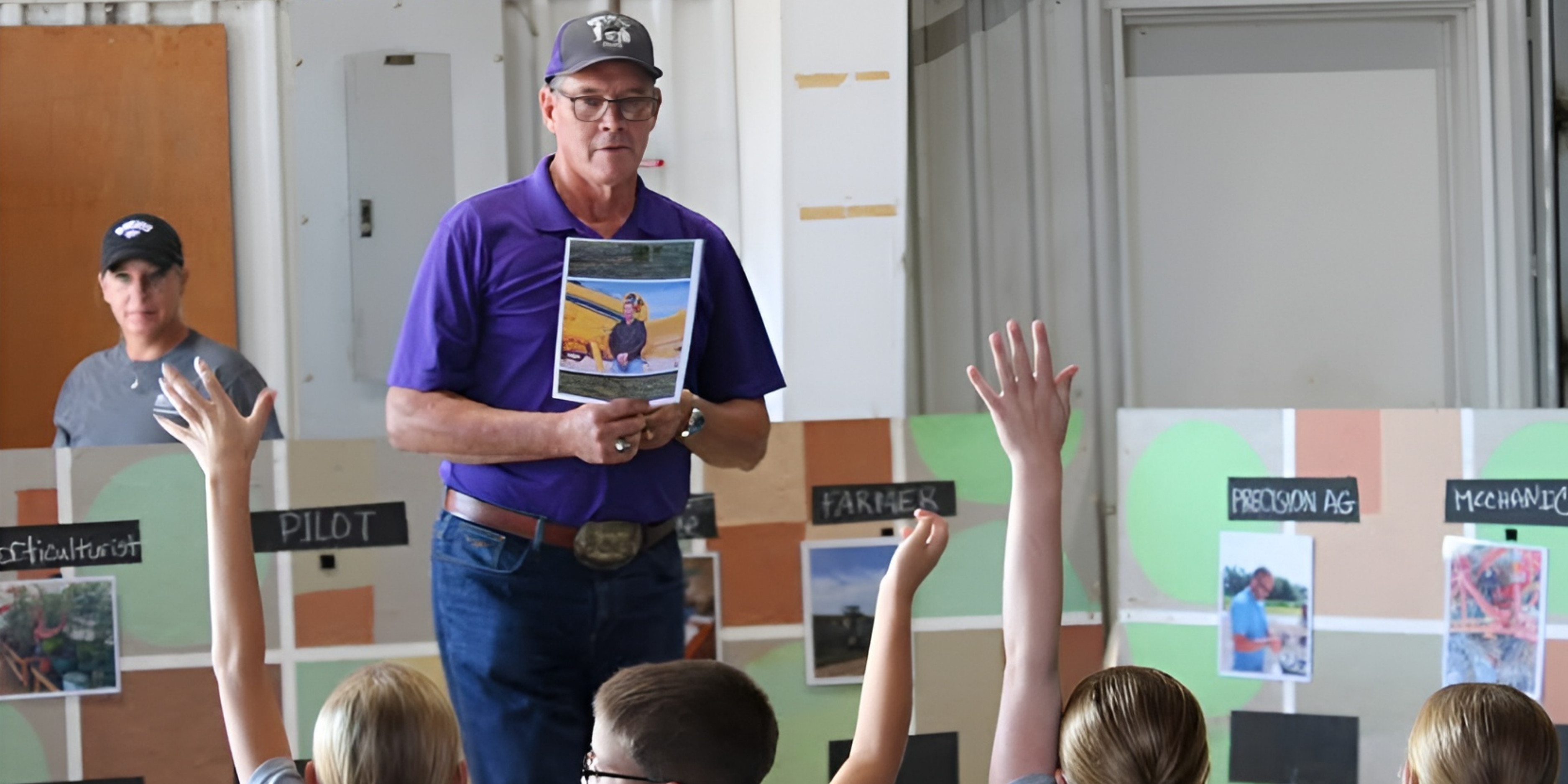 Jay Harris, in a purple shirt and hat, holds up a photo while speaking to a group of children with raised hands during a Kids Ag Day presentation.