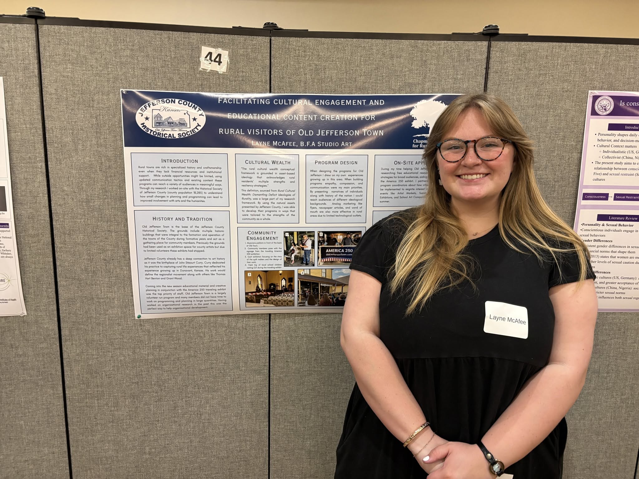 A female college student smiles and stands in front of a research poster in a college ballroom.