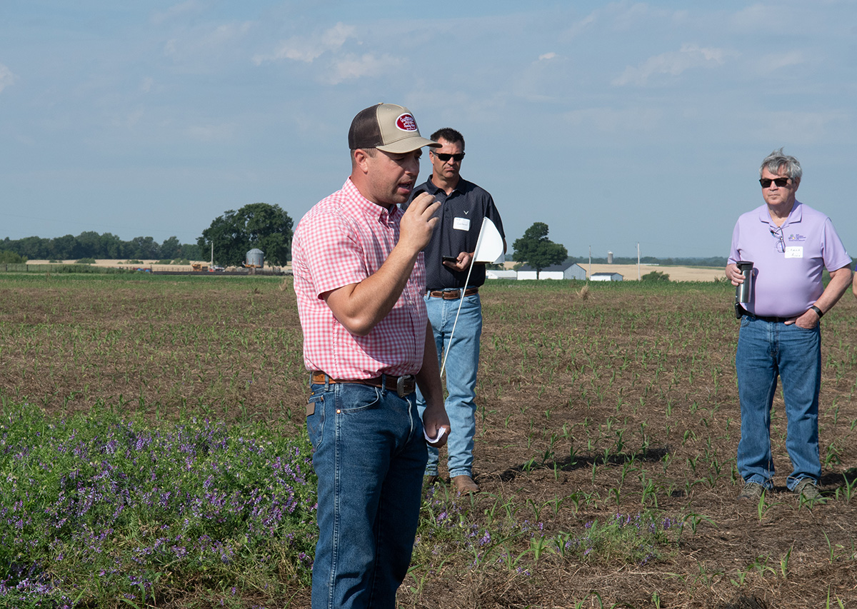 A man in jeans and a salmon checkered shirt speaks in a field with two men standing behind him.