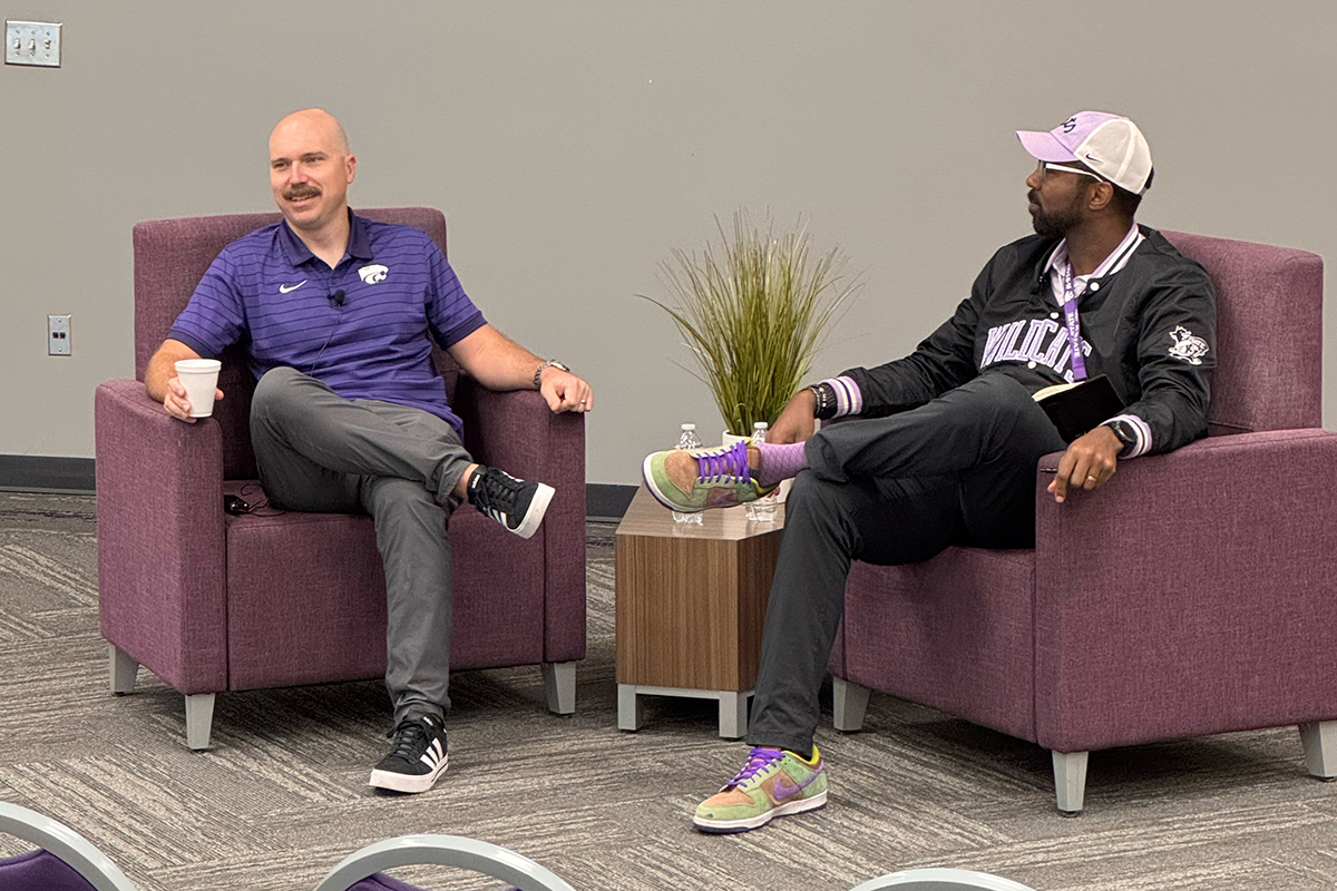 A mustached man in a purple polo has a moderated discussion at the front of an auditorium with a college professor.