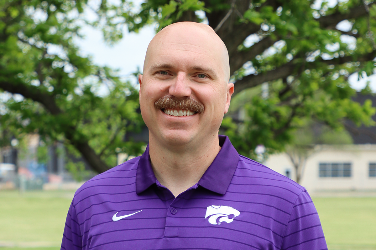 A mustached man in a purple shirt smiles for a portrait.