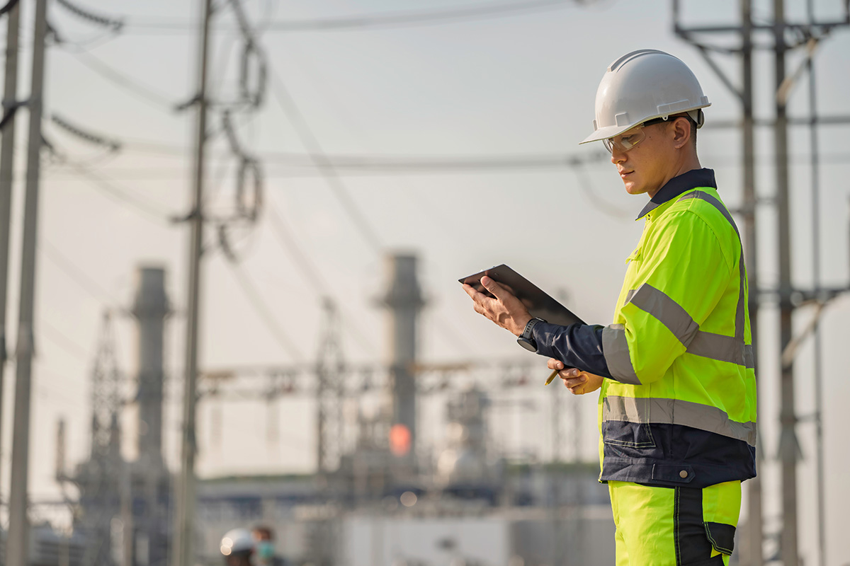 A man in a white hard hat and a yellow vest stands in the foreground. In the background is a large power plant.