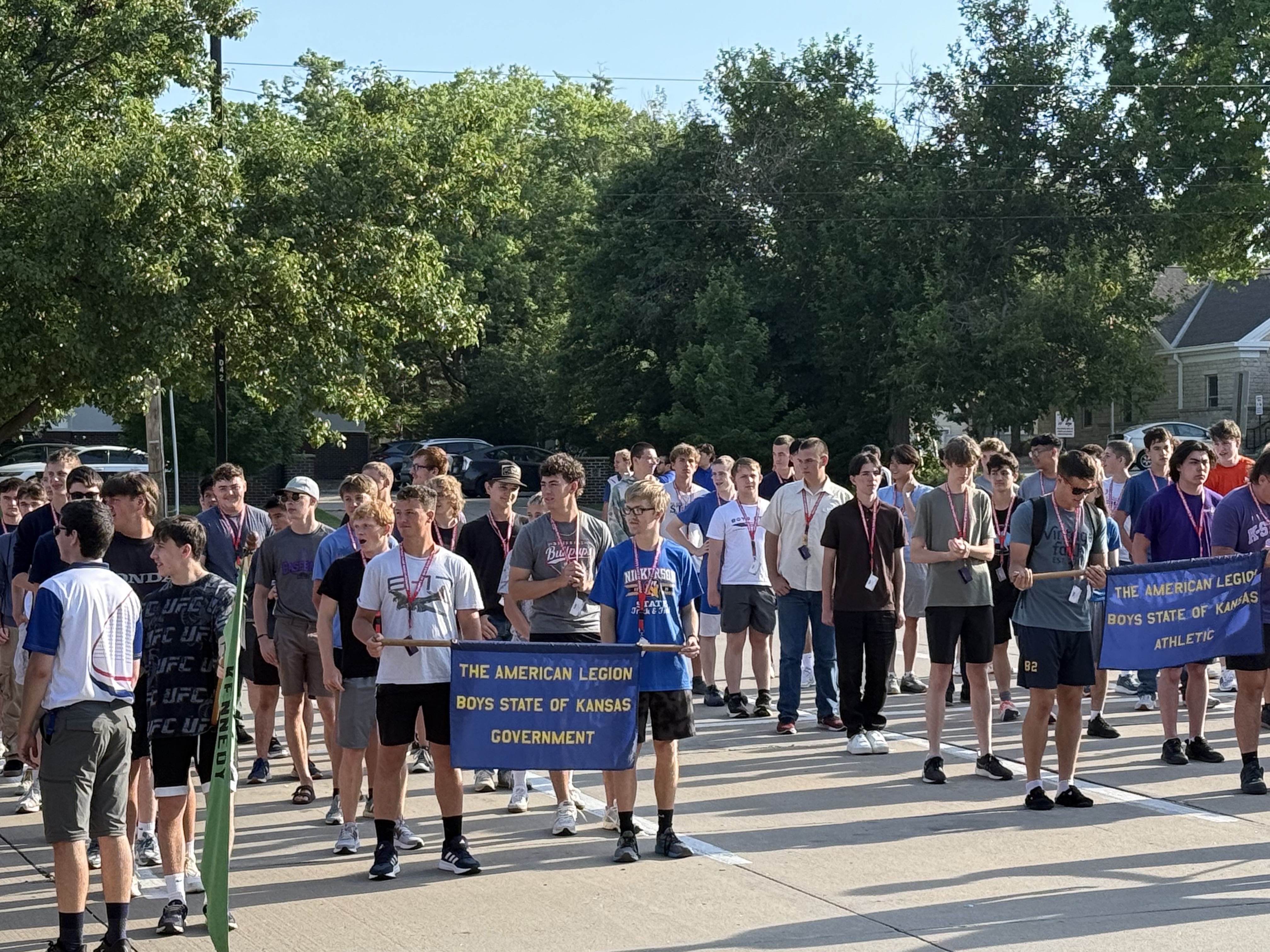 A group of high-school aged boys line up in a parking lot and hold up banners as part of a summer camp.