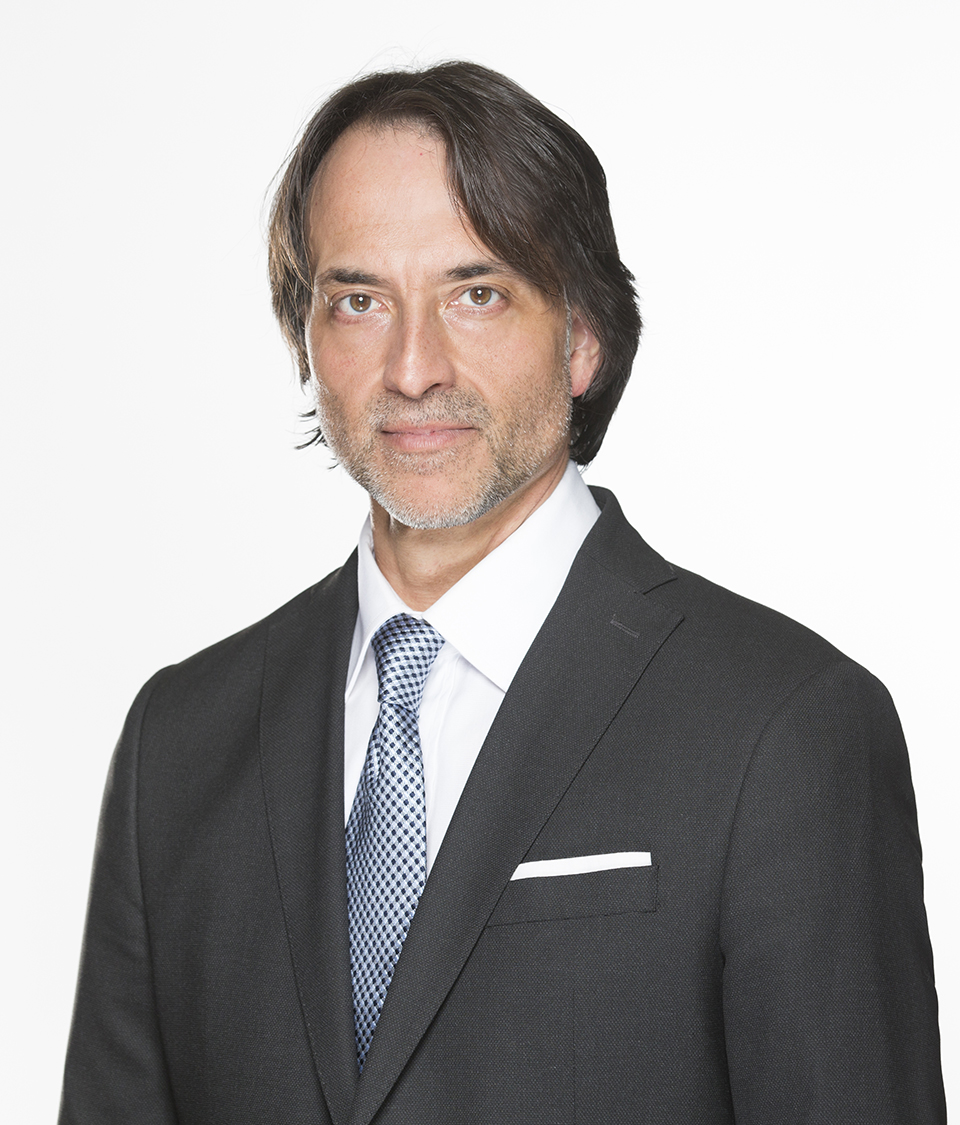 A man with dark hair and a gray suit with a blue checkered tie is pictured against a white background.