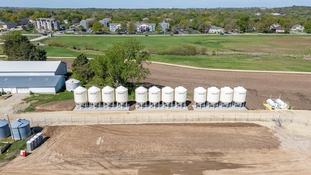 A row of twelve white agricultural hopper bins stands in a field with green grass visible in the background and some housing and cars.