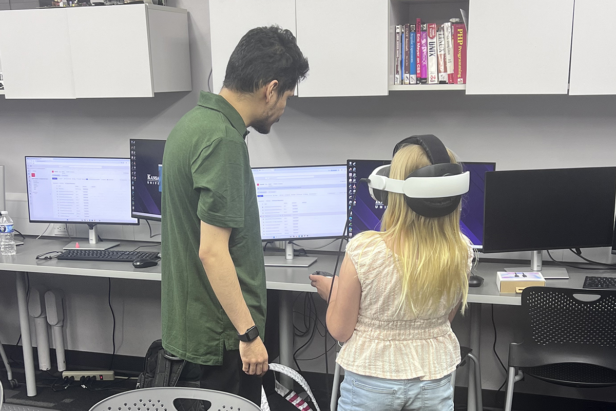 A college student interacts with a young girl as she wears a virtual reality headset and attempts to play a game.